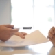 Person Handing Personal Loan Documents and Pen To Another Over a White Table